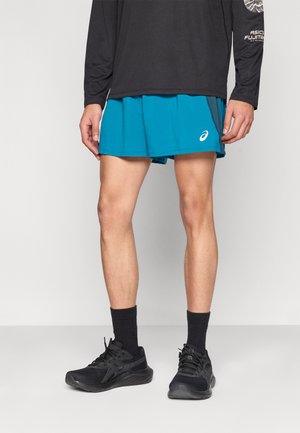 Türkiz atlétikai rövidnadrág rugalmas derékszíjjal, hosszú ujjú fekete felsővel párosítva. Fekete sportcipő sötét zoknival egészíti ki a szettet.