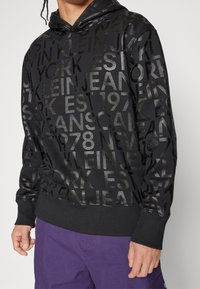 Black hoodie featuring a textured print of various brand names and dates in a glossy finish. Includes a drawstring hood and ribbed cuffs.