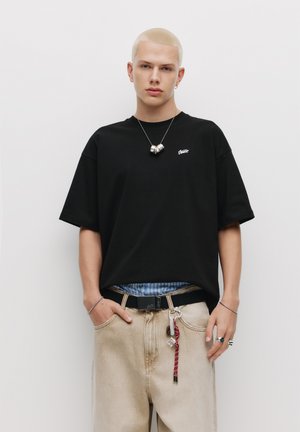 Schwarzes Baumwoll-T-Shirt mit kurzen Ärmeln und kleinem Logo, kombiniert mit hellbeigen Jeans und darüber getragen mit einem blauen karierten Hemd.