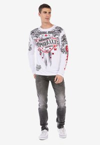 Langärmliges weißes Hemd mit schwarzen und roten Grafikdesigns, darunter florale Muster und ein Wappen-Logo. Kombiniert mit grauen Jeans und weißen Sneakern.