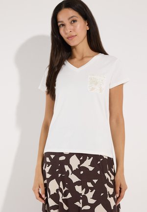 Frau trägt ein weißes V-Ausschnitt-T-Shirt mit kleinem Blumenmuster auf der Tasche und einen braunen Rock mit abstrakten beigen Mustern.