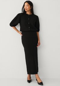 Schwarz zwei-teiliges Outfit mit einer kurzen, strukturierten Bluse mit Puffärmeln und einem hoch taillierten, bodenlangen Rock. Perlenähnliche Knöpfe setzen Akzente.