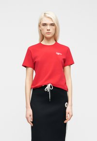 Νεαρή γυναίκα με ίσια ξανθά μαλλιά φοράει κόκκινο κοντομάνικο πουκάμισο και μαύρη φούστα με λευκό κορδόνι και λευκές ρίγες στο πλάι,  στέκεται μπροστά σε λευκό φόντο.