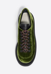Zapatilla de terciopelo verde con suela de goma negra, cordones marrones y ojetes de metal. Presenta una lengüeta acolchada y un acabado texturizado.