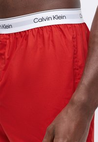 Rde plavalne kratke hlače z gladko teksturo in elastičnim pasom, ki na belem ozadju nosijo črno blagovno znamko "Calvin Klein".