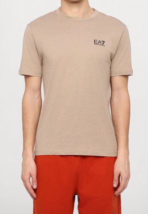 Mężczyzna ubrany w beżowy T-shirt EA7 Emporio Armani z krótkim rękawem i czerwone spodnie na jednolitym białym tle.