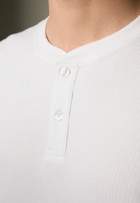 Camisa henley blanca de canalé con cuello bajo, que cuenta con dos botones blancos en la tapeta y una tela suave y texturizada.