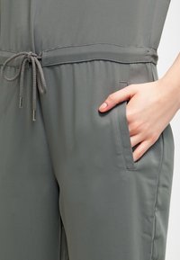 Szary kombinezon z paskiem ściąganym w talii, boczną kieszenią, gładką fakturą i luźnym krojem, wyróżniający się minimalistycznym designem.