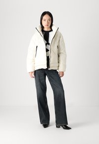 Krem krāsas pūkainā jaka ar augstu apkakli, melnu rāvējslēdzēju un sānu kabatām. Apvienota ar plašiem tumšas džinsa biksēm un melniem papēžiem.