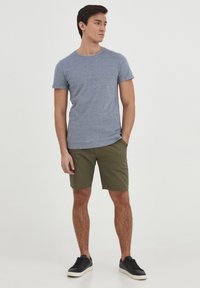 T-shirt gris à manches courtes associé à un short vert olive. Le modèle se tient les mains dans les poches, portant des baskets noires sur un fond uni.