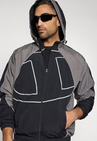 Chaqueta rompevientos negra y gris con acentos de contorno blanco, cierre de cremallera frontal, puños elásticos y capucha. Tejido ligero texturizado.