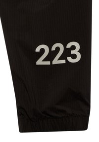 Černé sportovní kalhoty s proužkovanou texturou, na levé noze mají velké bílé číslo "223" a elastický manžet na spodním okraji.