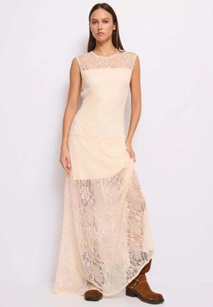 Donna che indossa un abito lungo senza maniche in pizzo beige abbinato a stivaletti in camoscio marrone, in piedi davanti a uno sfondo bianco.