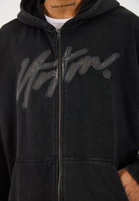 Black zip-up hoodie made of soft fabric, featuring a prominent glossy logo on the front. Includes a spacious hood and front pockets.
