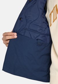 Marinblå pufferväst inuti med kviltad vaddering, tryckknappsstängning och en stor ficka i botten med en slät yta.