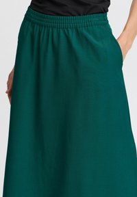 Jupe teal avec une ceinture élastique, confectionnée dans un tissu lisse, présentant une coupe décontractée et des poches latérales, longueur juste au-dessus du genou.