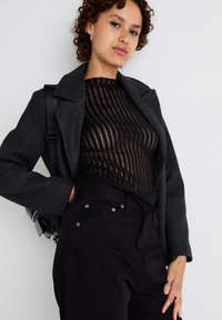 Top nero trasparente con strisce verticali, abbinato a un blazer grigio scuro su misura. Pantaloni neri con bottoni di ottone. Look casual.