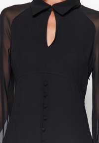 Schwarzes Kleid mit Kragen, Schlüsselloch-Ausschnitt und langen durchsichtigen Ärmeln. Glattes Material mit einer Knopfleiste auf der Vorderseite.