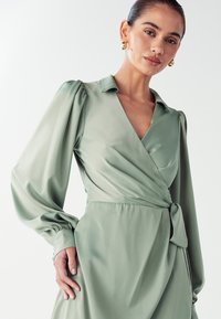 Zelené saténové šaty s hlbokým výstrihom do V, dlhými puff rukávmi a detailom viazania v páse, s hladkou a jemnou textúrou.