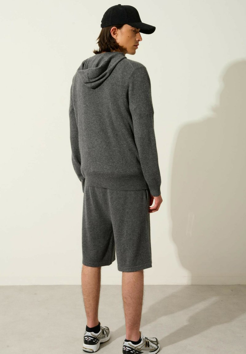 Szary, dzianinowy hoodie z kapturem, zestawiony z szarymi szortami. Model nosi czapkę w kolorze czarnym i sneakersy, prezentując nowoczesny, fakturowany design.