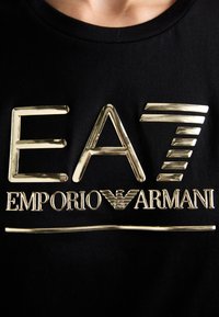 Crna majica s otisnutim zlatnim logotipom "EA7 EMPORIO ARMANI" koja ima jedinstvenu teksturiranu završnu obradu i elegantan dizajn sprijeda.