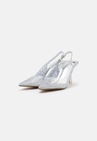 Sudraba siksnē vairogto papēžu kurpes ar norādītu pirkstu dizainu, dekorētas ar rhinestones un caurspīdīgu bloka papēdi. Gluda, spīdīga tekstūra.