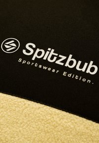 Tessuto in pile nero e beige con una cucitura. Testo "Spitzbub Sportswear Edition" in bianco. Texture liscia e colori a contrasto.