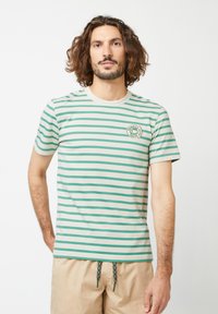 Gestreiftes T-Shirt in hellbeige mit grünen horizontalen Streifen. Hat einen runden Ausschnitt und einen kreisförmigen Logo-Patch auf der Brust.