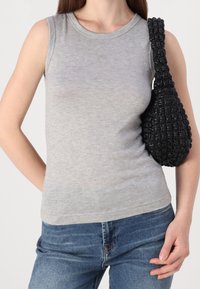 Top grigio senza maniche con scollatura rotonda e un piccolo adornamento triangolare, abbinato a una borsa nera con texture. Jeans blu visibili.