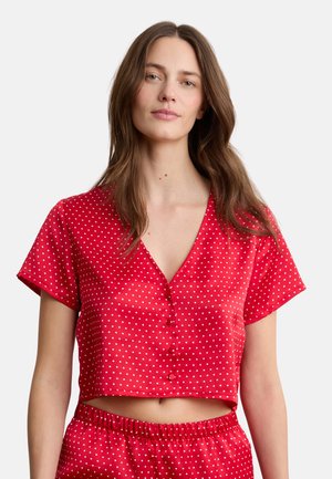 Jonge vrouw die een rood crop top met korte mouwen draagt met witte polka dots en bijpassende hoge taille broek tegen een witte achtergrond.