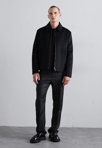 Black jacket with a zip closure and collar, paired with black fitted pants and chunky black shoes, against a neutral background.