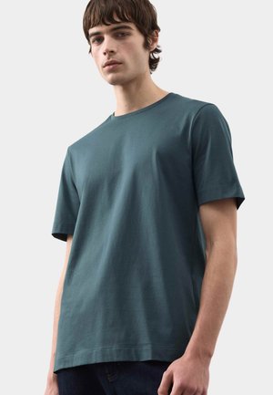 Giovane uomo che indossa una semplice t-shirt a maniche corte color verde petrolio scuro, in piedi davanti a uno sfondo grigio chiaro.