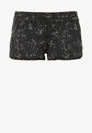 Femme debout portant un soutien-gorge de sport noir, un short sportif noir à motifs, et des chaussures de running noires et violettes sur fond blanc.