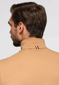 Beige Poloshirt mit hohem Kragen, ausgestattet mit einem kleinen blau-weiß-roten Etikett auf der Rückseite. Glatte Textur und taillierte Passform.