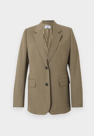 Zwarte vrouw die een beige blazer, wit blouse met knopen, donkerbruine broek en zwarte hakken draagt, staand met één hand in de zak.