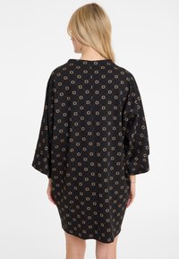 Zwarte stoffen jurk met een geometrisch patroon in gouden accenten, losse pasvorm, driekwart mouwen en een ronde halslijn.