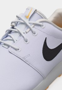 Biela kožená teniska s čiernym logom Nike Swoosh, bielymi šnúrkami a malým žltým logom Nike na jazyku, detailný záber.