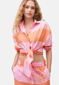Camicia collared e annodata in tessuto tie-dye rosa e arancione. Maniche corte e vestibilità rilassata, abbinata a pantaloni corti con coulisse annodati dello stesso tessuto.