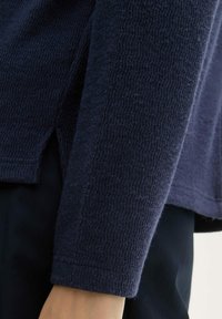 Pull en maille bleu marine texturé avec un tissu doux et tissé. Détails incluant un ourlet légèrement arrondi et des manches longues avec une coupe décontractée.