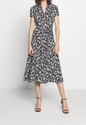 Zwarte bloemenjurk met korte mouwen, getailleerde taille en een uitlopende rok, bedrukt met een wit bloemenpatroon. Draagt zwarte sandalen met hak.