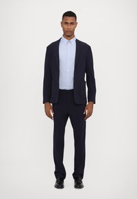 Costume bleu marine ajusté avec deux poches avant, porté sur une chemise à boutons bleu clair. Des chaussures noires complètent la tenue formelle.