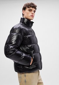 Μαύρο μπουφάν πούφ με γυαλιστερή επιφάνεια, διαθέτοντας ψηλό γιακά, ραμμένες πάνελ και τσέπες με φερμουάρ στα πλάγια.