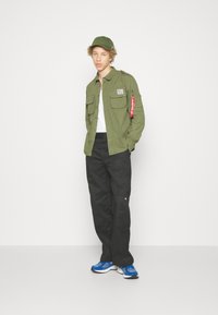 Olive green long-sleeve shirt with button-up front, two chest pockets, and a red tag. Black cargo pants and blue athletic shoes complete the look.
