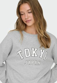 Ung kvinde med langt lysebrunt hår, iført en grå sweatshirt med "TOKYO JAPAN" trykt i hvide blokbogstaver.