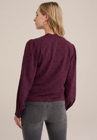 Bordeauxfarbiges Langarmoberteil mit feiner Textur und puffigen Ärmeln, kombiniert mit grauen Jeans mit Gesäßtaschen und Nähdetails.