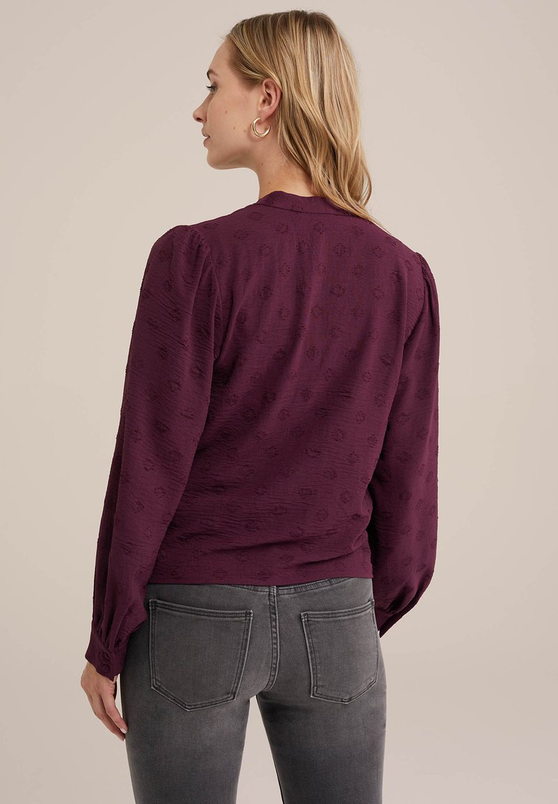 Bordeaux langarmtop met subtiele textuur en pofmouwen, gecombineerd met grijze jeans met achterzakken en stikdetails.