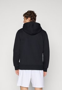 Sweat-shirt noir avec capuche, présentant une texture lisse et des poignets et un bord ribbelés, affiché de dos avec un short blanc.