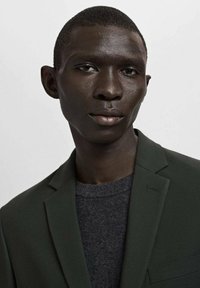 Dunkelgrüner Blazer mit einem strukturierten Design, einem Revers mit Kerbkragen und einem einzelnen Knopf, getragen über einem dunkelgrauen Pullover. Glatte Textur und maßgeschneiderte Passform.