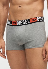 Torse masculin portant un boxer gris Diesel avec une ceinture élastique noire comportant le logo Diesel blanc et orange.