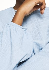 Handen vilar nära ansiktet, iklädd en ljusblå texturerad långärmad topp och ett guldhalsband synligt vid halslinjen.